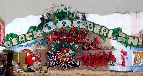 Mann auf einer Leiter bemalt eine Hauswand, Schriftzug "Beach Bar Berlin"
