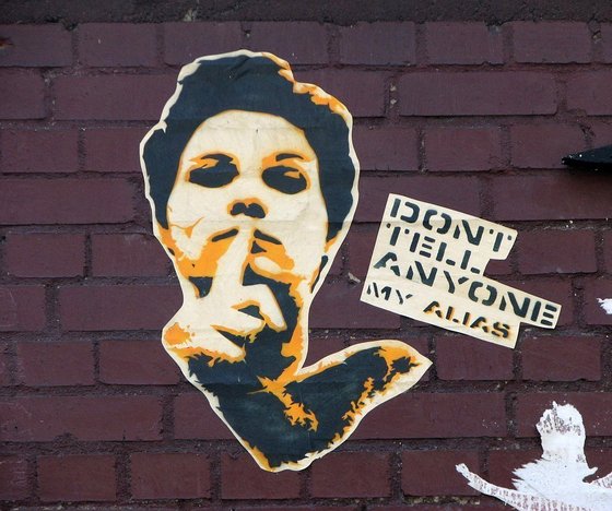 Portrait eines Mannes mit Finger vor dem Mund, Schriftzug "Don't tell anyone my Alias"